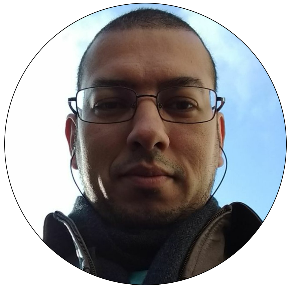 Foto redonda contemplando apenas o meu rosto. O fundo é um céu azulado. Uso óculos e fones de ouvido. Cachecol em tom cinza escuro e blusa de couro marrom. Possuo barba semi-serrada e cabelo bem curto. Olhos castanhos escuros e olhar fixo para a câmera.